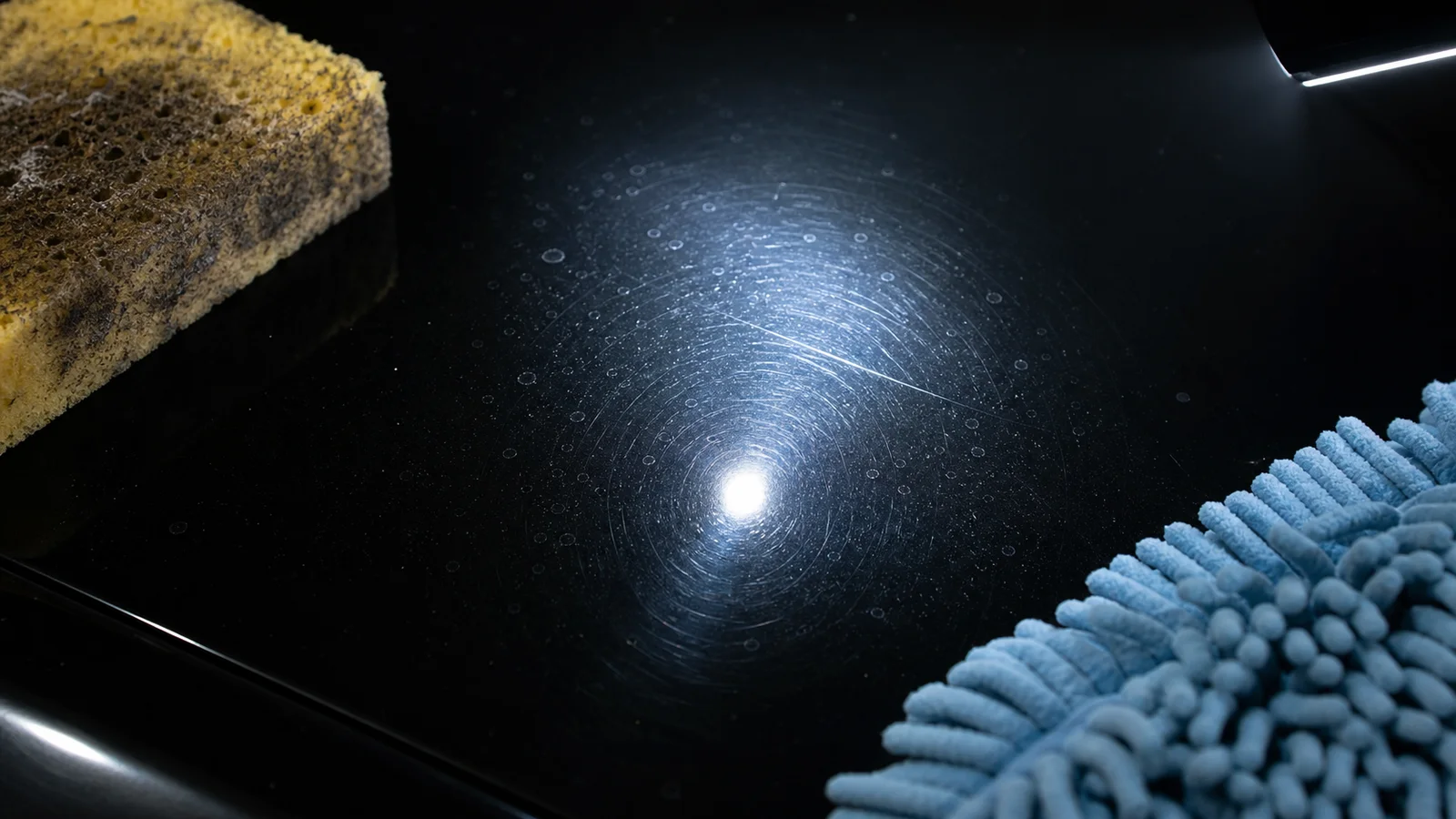 Inspection light revealing swirl marks, water spots, and a light scratch beside a dirty sponge and clean wash mitt